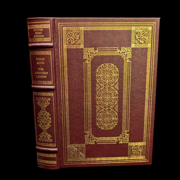 A signed first edition hardcover book titled 'The Anatomy Lesson' by Philip Roth, featuring a maroon and gold cover with intricate designs.