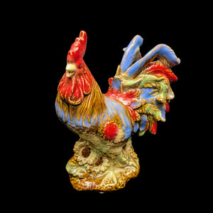 A hand-painted ceramic rooster figurine with a red comb and blue and green feathers, positioned on a beige base.