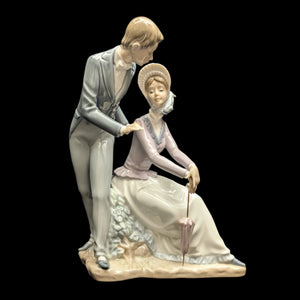 A Lladro figurine of a romantic couple, with the man standing and the woman sitting, holding a umbrella. The woman is wearing a dress and a hat, and the man is in a suit.