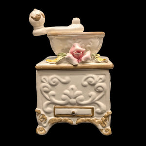 A vintage Capodimonte porcelain coffee grinder figurine, featuring intricate floral patterns and a pink flower detail on the top, placed against a black background.