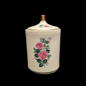 A vintage German porcelain jar with a lid, featuring a pink rose motif on the front, with green leaves and a multicolored floral pattern.